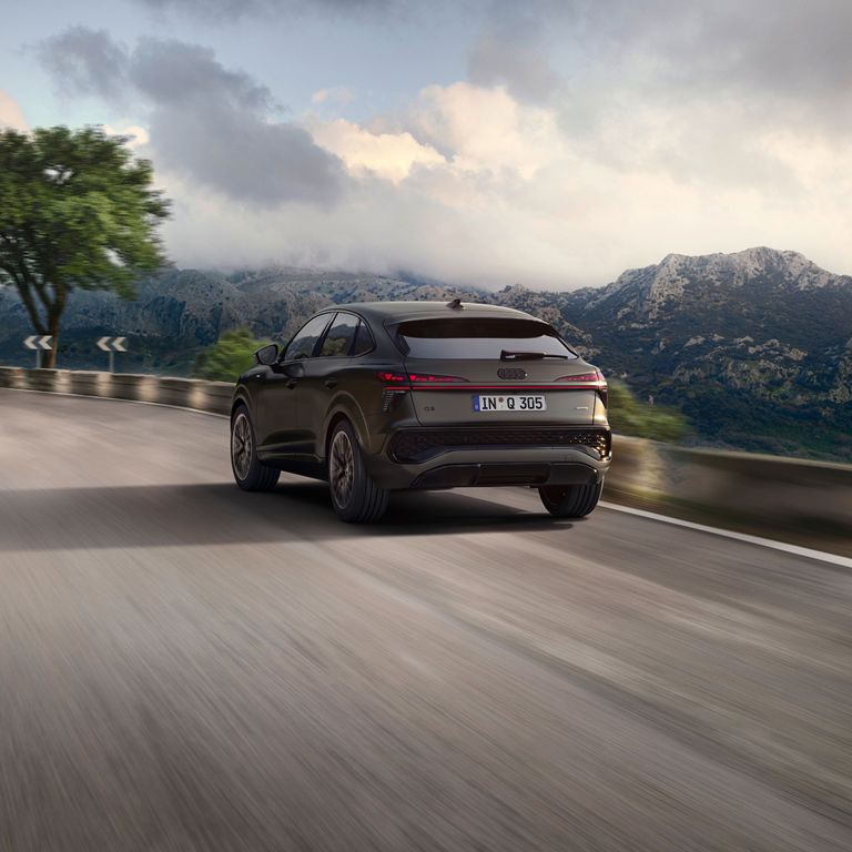 A matte black Audi Q3 driving on a winding mountain road with scenic cliffs and a cloudy sky behind.