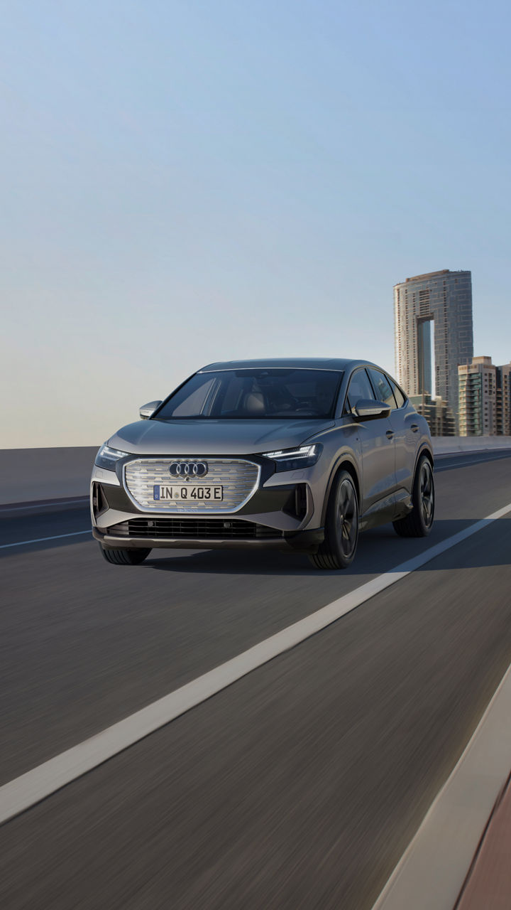 A grey Audi Q4 Sportback e-tron driving on a modern highway with a city skyline in the background against a clear sky.