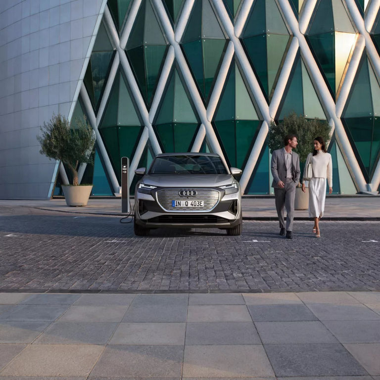An Audi Q4 Sportback e-tron parked at a charging station with modern architecture in the background, as a couple strolls by.