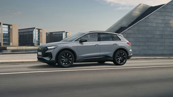 A grey Audi Q4 e-tron driving along a modern city street with contemporary buildings in the background under a clear sky.
