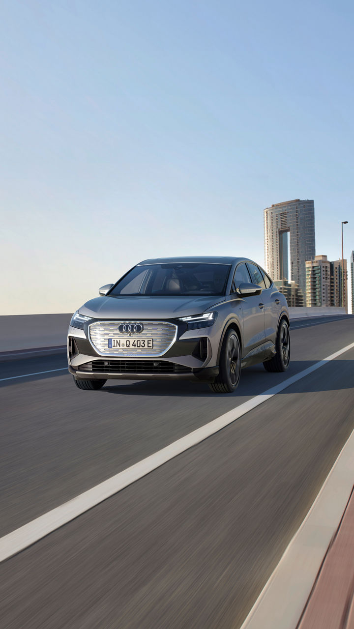 A grey Audi Q4 Sportback e-tron driving on a modern highway with a city skyline in the background against a clear sky.