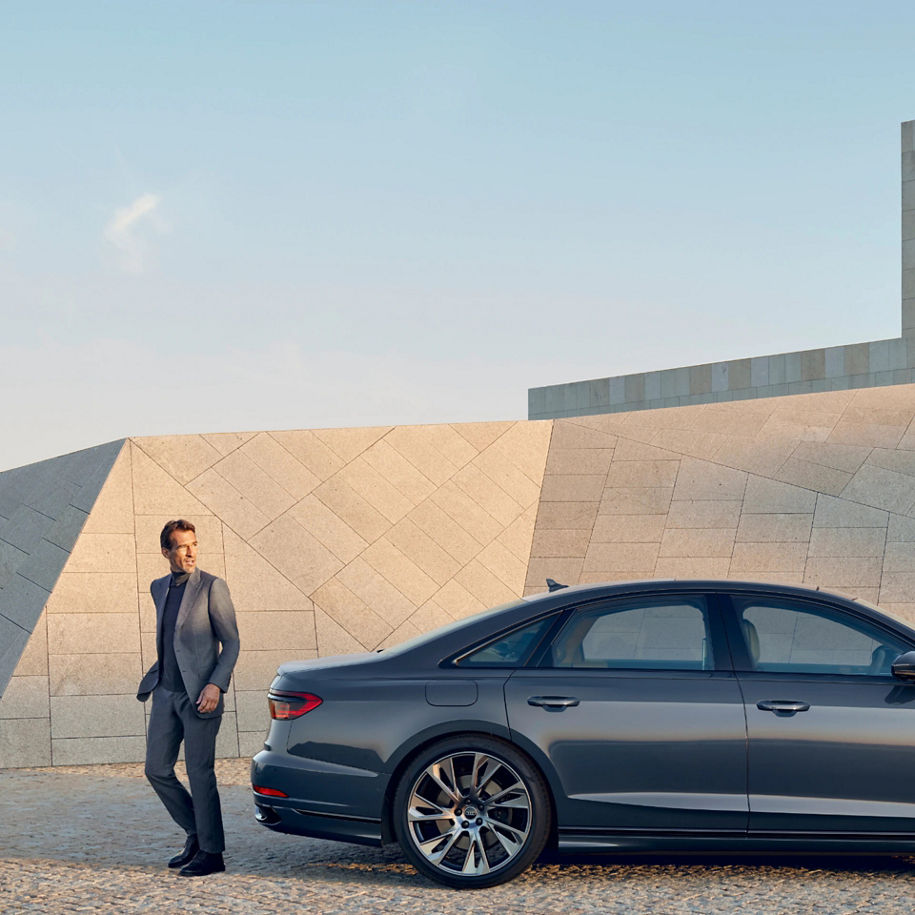 A person in a suit standing beside a sedan in front of a modern architectural structure.