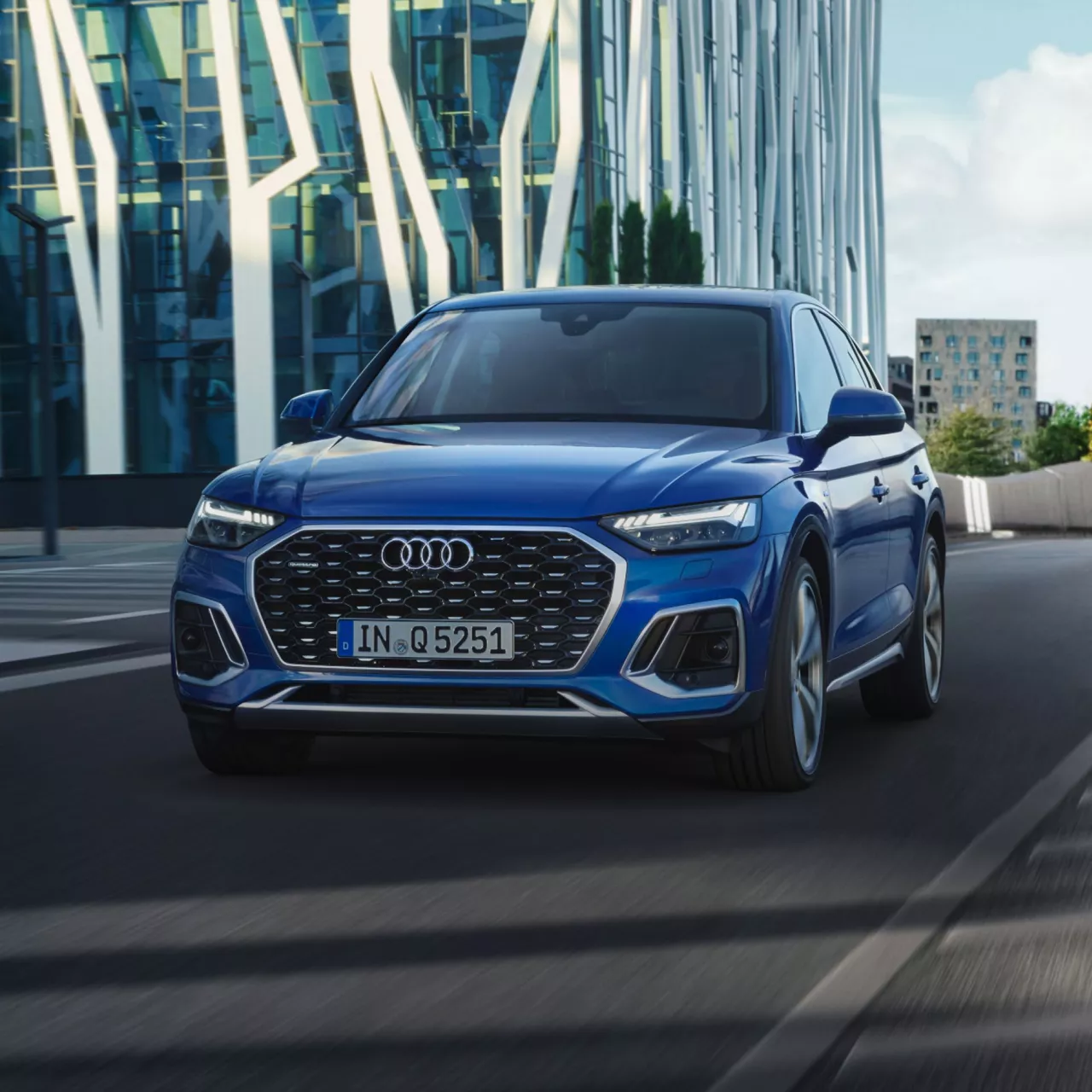 Blue Audi SUV driving through a city road, with a modern glass building in the background.