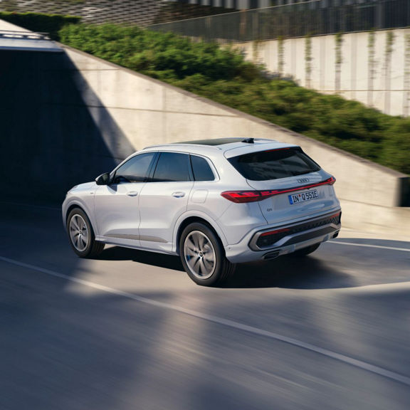 A sleek white Audi Q5 SUV e-hybrid drives dynamically on a modern road, with greenery in the background and a tunnel entrance ahead.