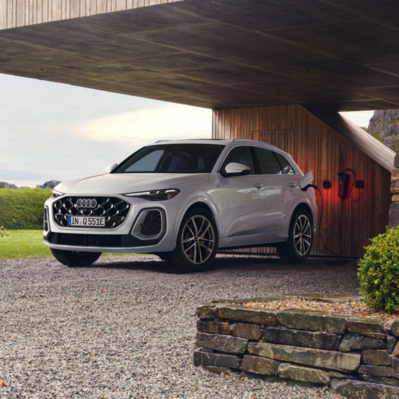 A sleek Audi Q5 e-hybrid parked under a modern shelter, plugged into an electric charger, surrounded by greenery and stone accents.
