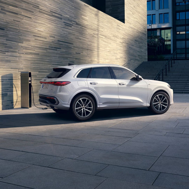 White SUV parked next to a charging station, set against a modern stone wall and urban architecture in the background.