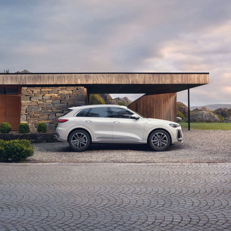 A white SUV parked beside a modern stone and wood house, set against a scenic backdrop of hills and clouds.