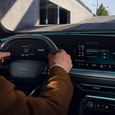 A driver grips the steering wheel of an Audi, with a sleek dashboard displaying digital controls and driving modes in a modern setting.