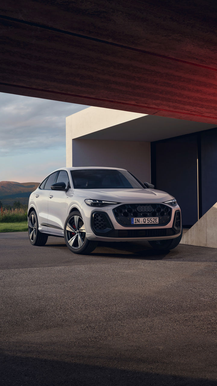 A white Audi Q5 Sportback e-hybrid with a bold honeycomb grille parked near a modern building, framed by distant mountains in the background.