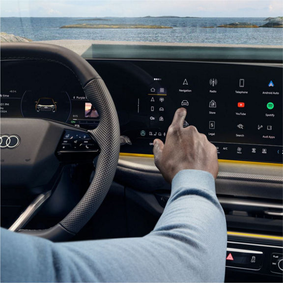 A close-up of a person using a touchscreen display in an Audi car, showcasing navigation, apps, and vehicle controls with a scenic ocean view.