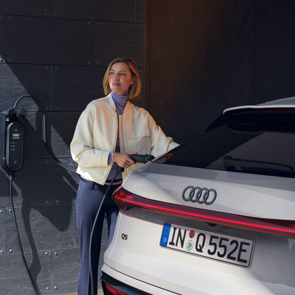 A woman in a cream jacket plugs a charging cable into a white Audi Q5 parked near a charging station. Modern garage setting.