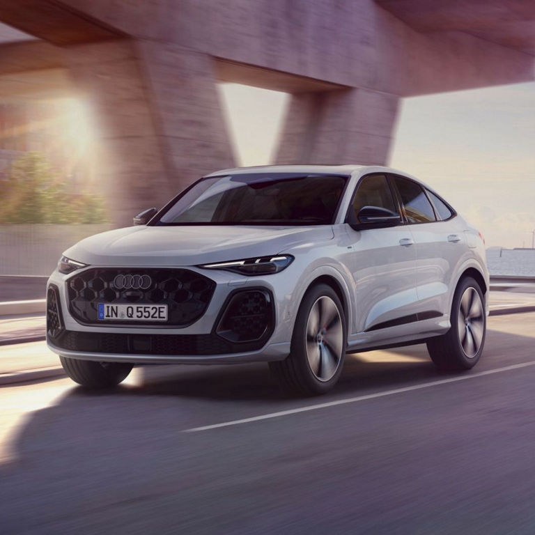 A white Audi Q5 Sportback e-hybrid drives on a scenic road under a bridge, with modern buildings and wind turbines in the background.