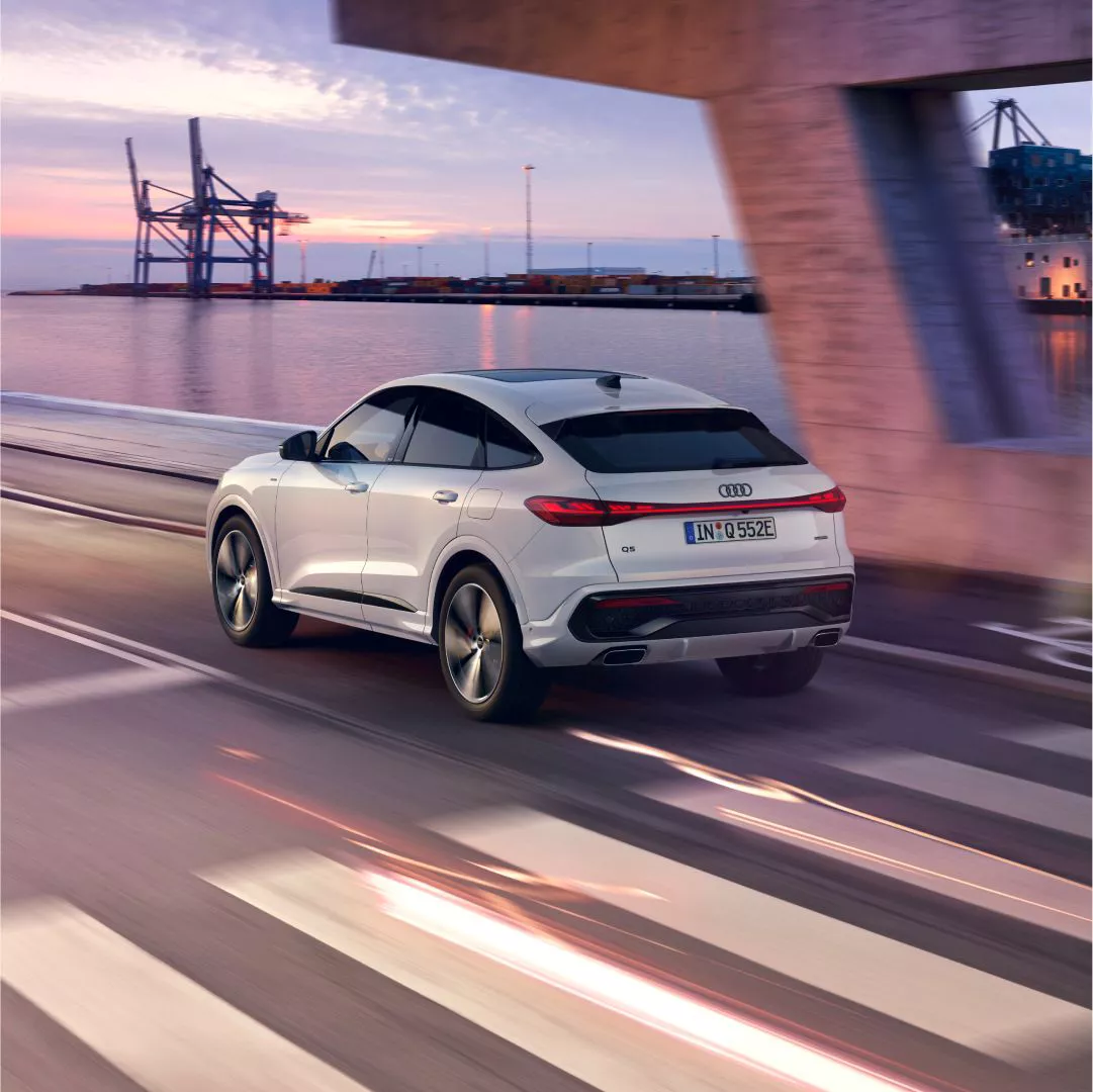 A white Audi Q5 Sportback e-hybrid drives along a waterfront road at sunset, with industrial cranes and shipping containers in the background.