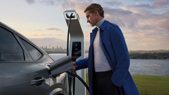 A person in a blue coat connects a charging cable to an electric vehicle at a modern charging station, with a city skyline in the background.