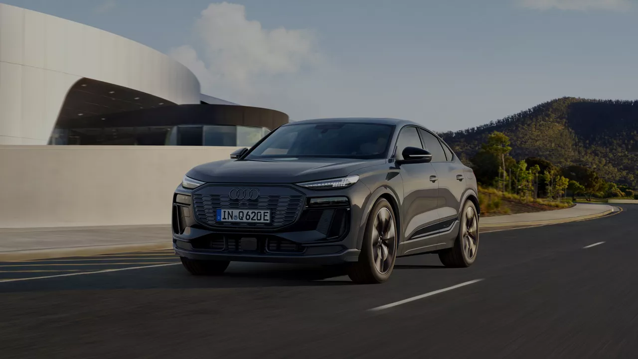 A sleek black Audi Q6 Sportback e-tron drives on a scenic road, modern architecture and hills visible in the background under a clear sky.