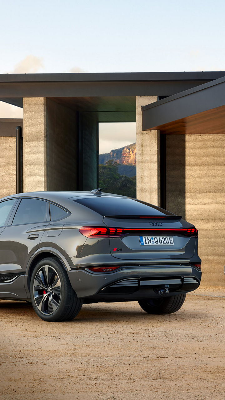 A sleek grey Audi Q6 Sportback e-tron parked in a stylish outdoor setting, with modern architecture and a boat trailer visible in the background.