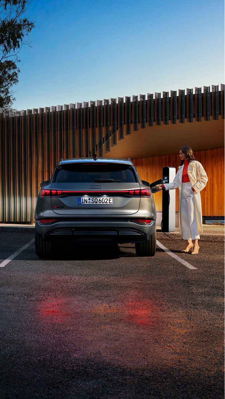 A woman in stylish attire plugs in an Audi SQ6 e-tron to a charging station, set against a sleek wooden structure.
