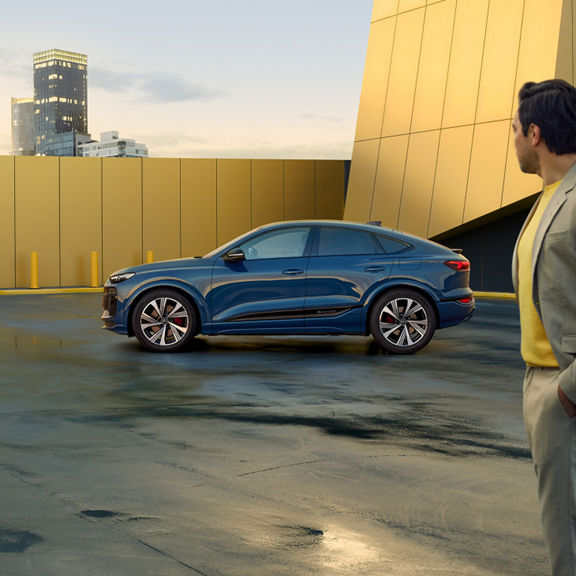 A man in a suite looking at a stationary blue SQ6 Sportback e-tron with a yellow building in the back