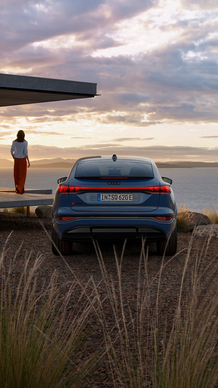 A blue Audi SQ6 Sportback e-tron seen from behind while park near a cliff with lake view at the back and woman standing on a house porch