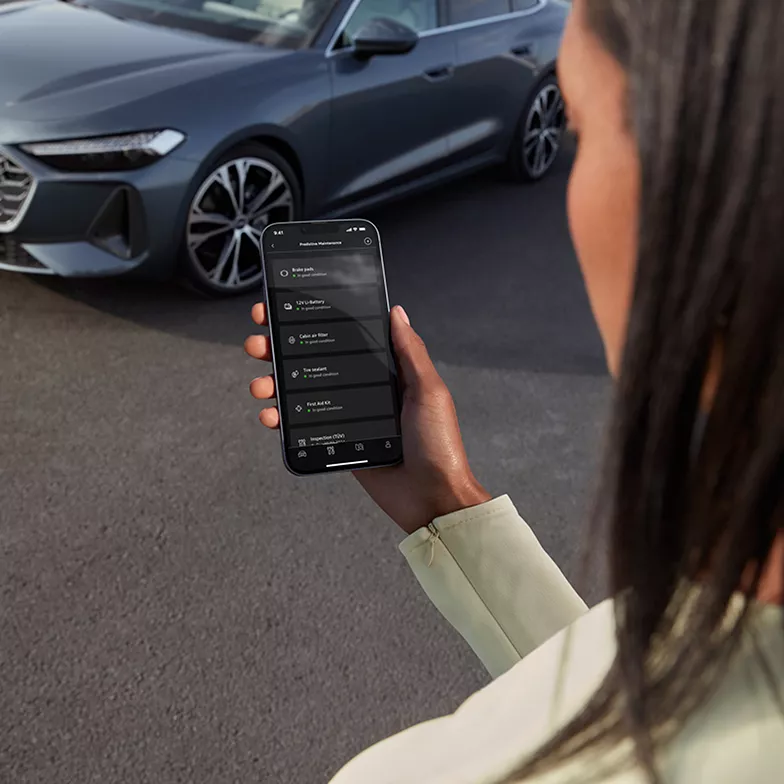 A woman using a smartphone to access car features, with a sleek gray vehicle parked in the background.