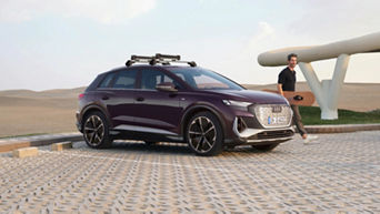 A man with a snowboard under his right arm and walking behind a parked Audi vehicle 