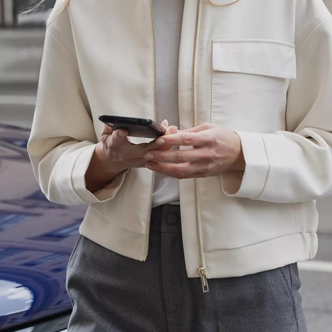 A person in a cream bomber jacket and gray pants stands outside, engaged with their smartphone.