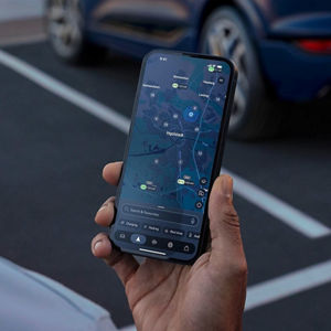 A hand holds a smartphone displaying a city map. The screen shows multiple green markers with labels, suggesting various locations or points of interest. The setting is casual, with a neutral background, conveying a sense of navigation and exploration.