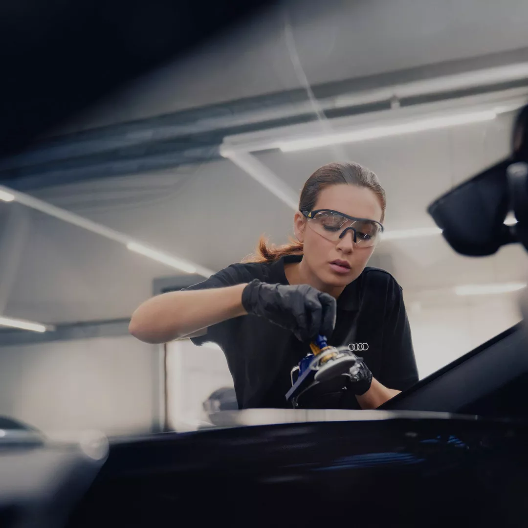 A woman with glasses is working on a car's engine, surrounded by tools in a workshop. 