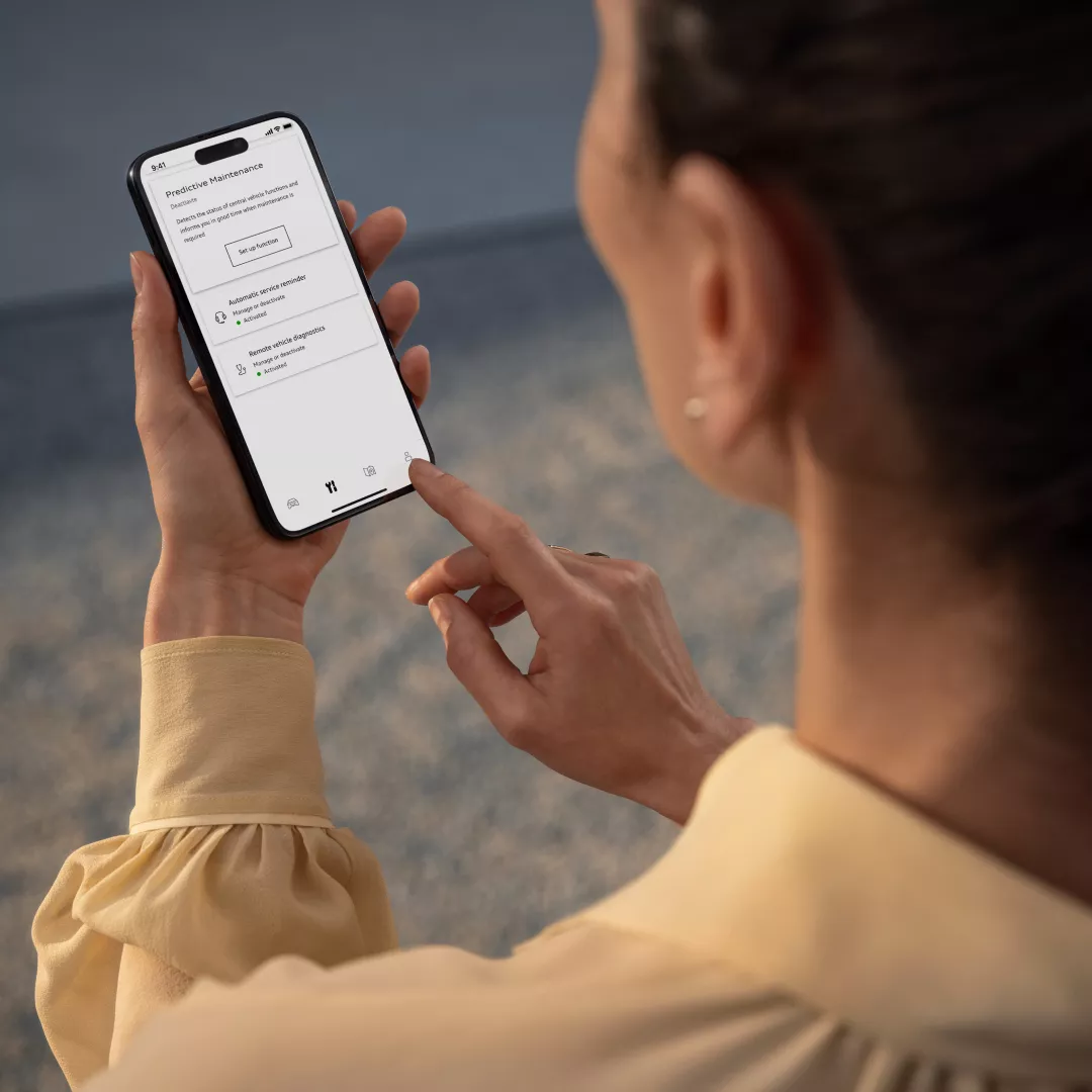 A woman in a yellow blouse holds a smartphone displaying a predictive maintenance app interface. She is tapping the screen, focused.