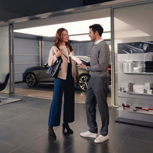 A woman in a stylish outfit interacts with a salesman inside a modern car showroom, showcasing cars and products on display.
