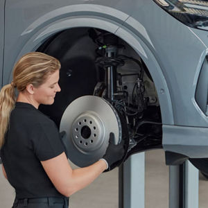 A technician in black attire is holding a brake disc while inspecting the wheel assembly of a car lifted for maintenance.