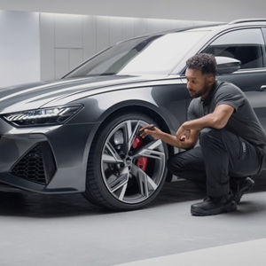 A mechanic inspects the wheel and brake system of a sleek grey Audi sports car in a modern, well-lit garage.
