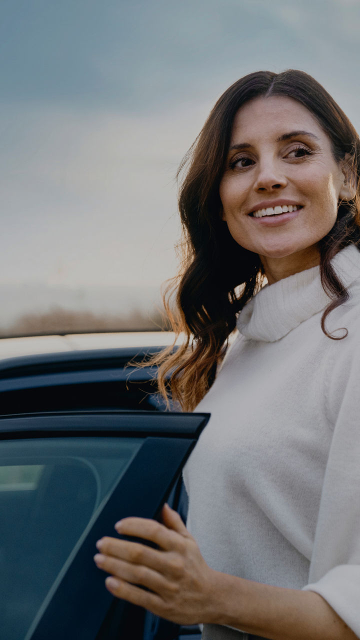 A woman stands beside her car, smiling and enjoying a sunny day outdoors.