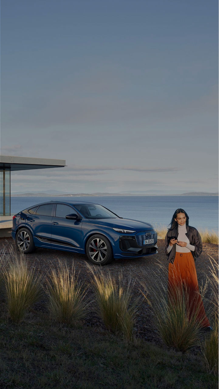 A woman stands using her phone near a blue electric Audi SUV parked by a modern house and charging station, with a scenic coastal view in the background.