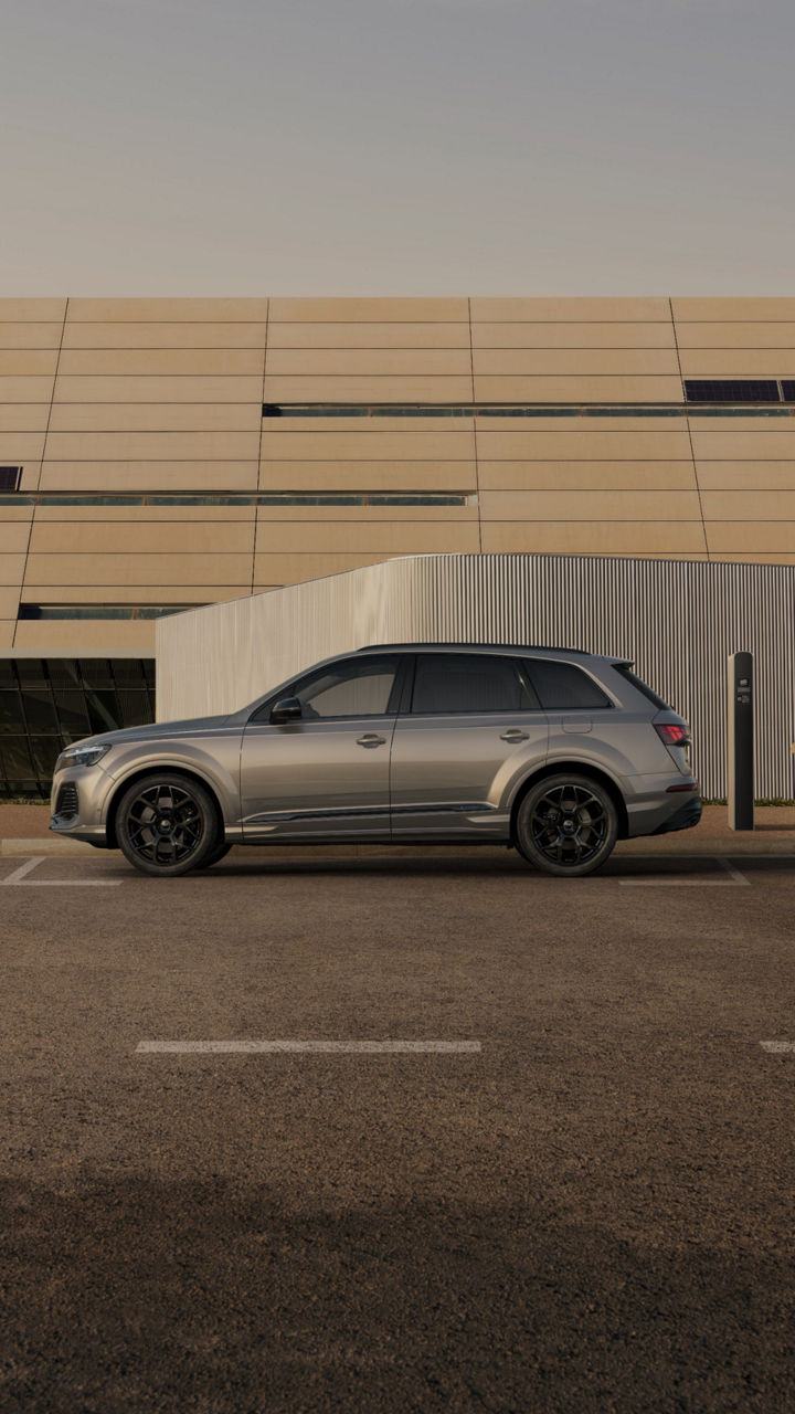 A sleek silver SUV parked beside a modern building, with a pedestrian approaching a charging station nearby.