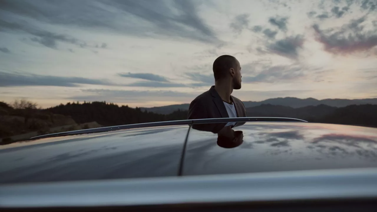 A man in a suit stands beside a car, gazing at a colorful sunset over mountains and a cloudy sky. Reflection visible on the car's roof.