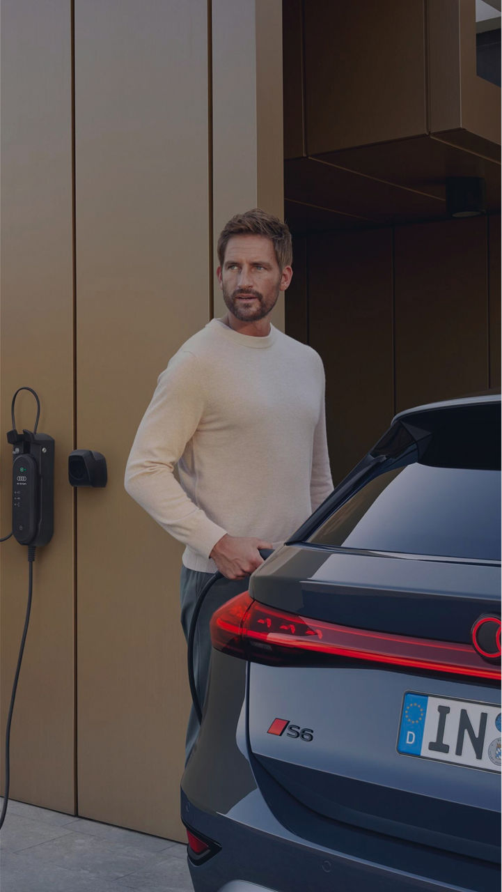 A man standing next to an Audi car with red light details and an EV charger on the wall behind
