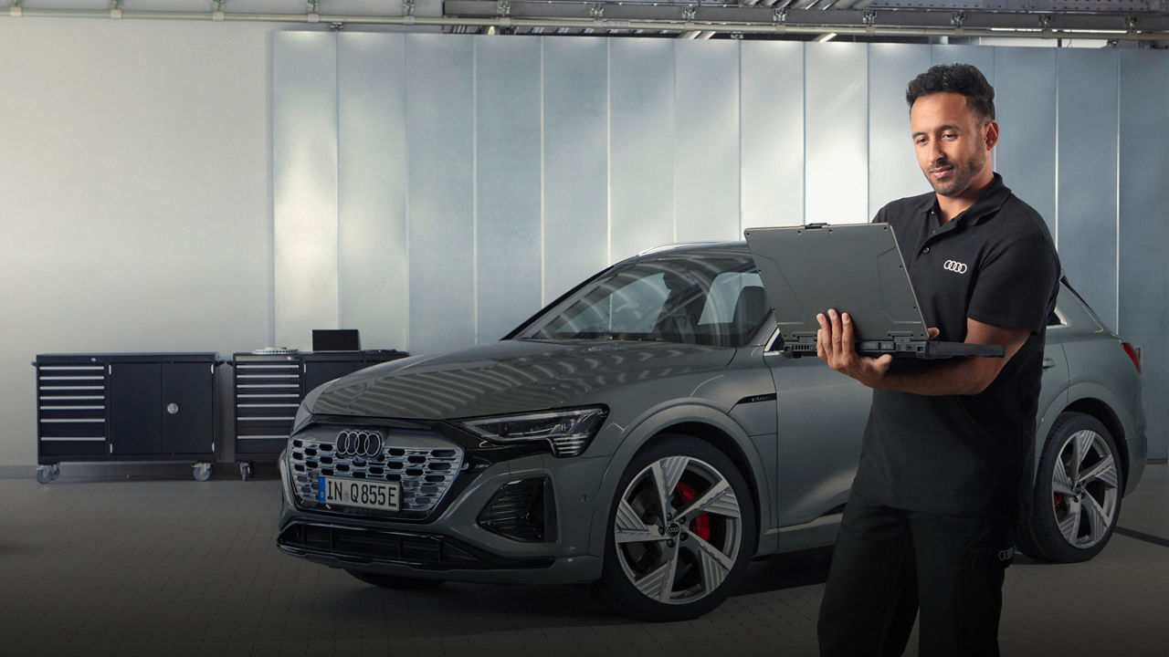An Audi technician looking at a laptop with an Audi vehicle parked in the background