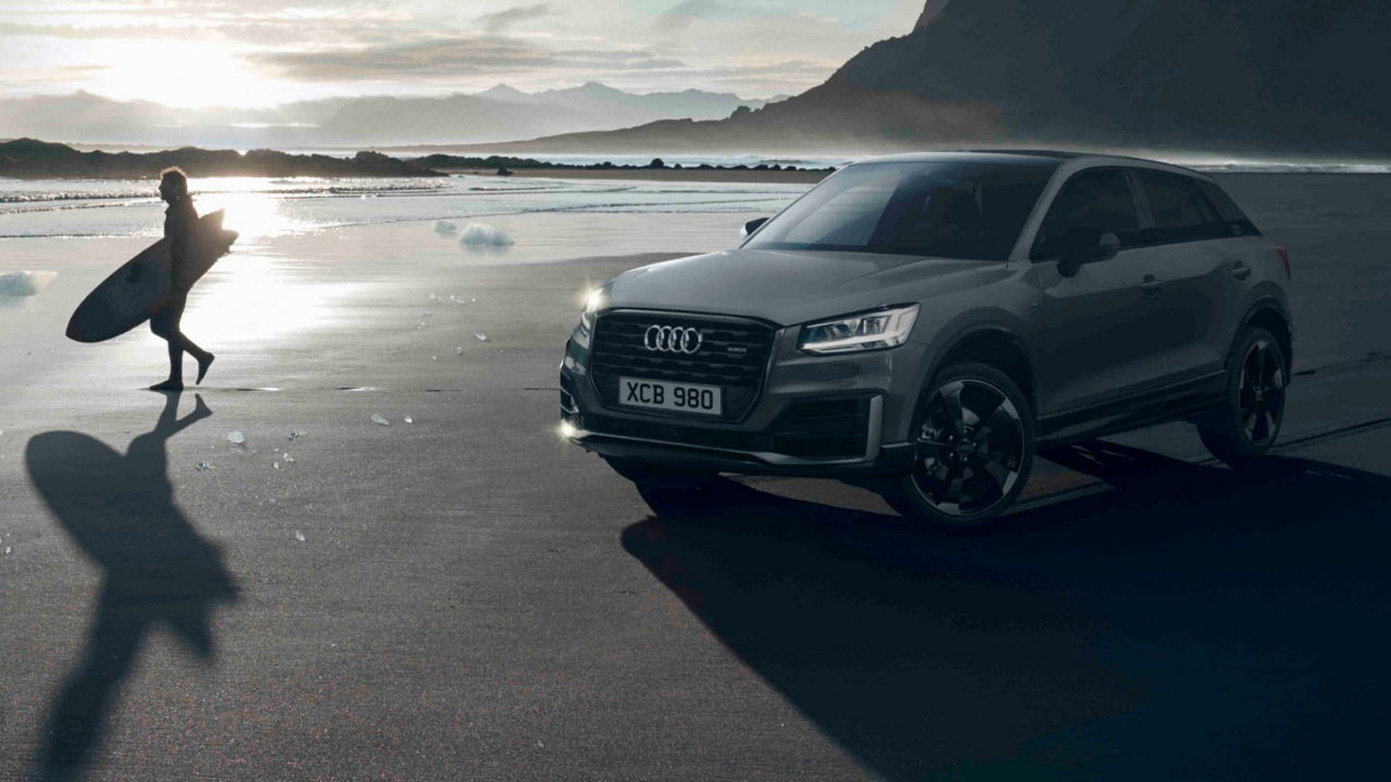 An Audi on a beach with a surfer walking by