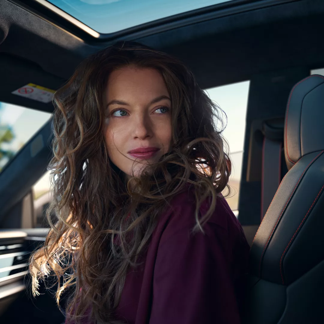 A woman with wavy hair sits in a car, wearing a burgundy top. Sunlight streams through the roof, illuminating the interior.