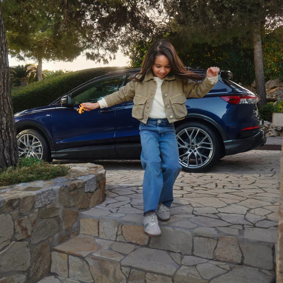 A child in a beige jacket and blue jeans descends stone steps, holding a toy, with a blue car parked nearby and trees in the background.