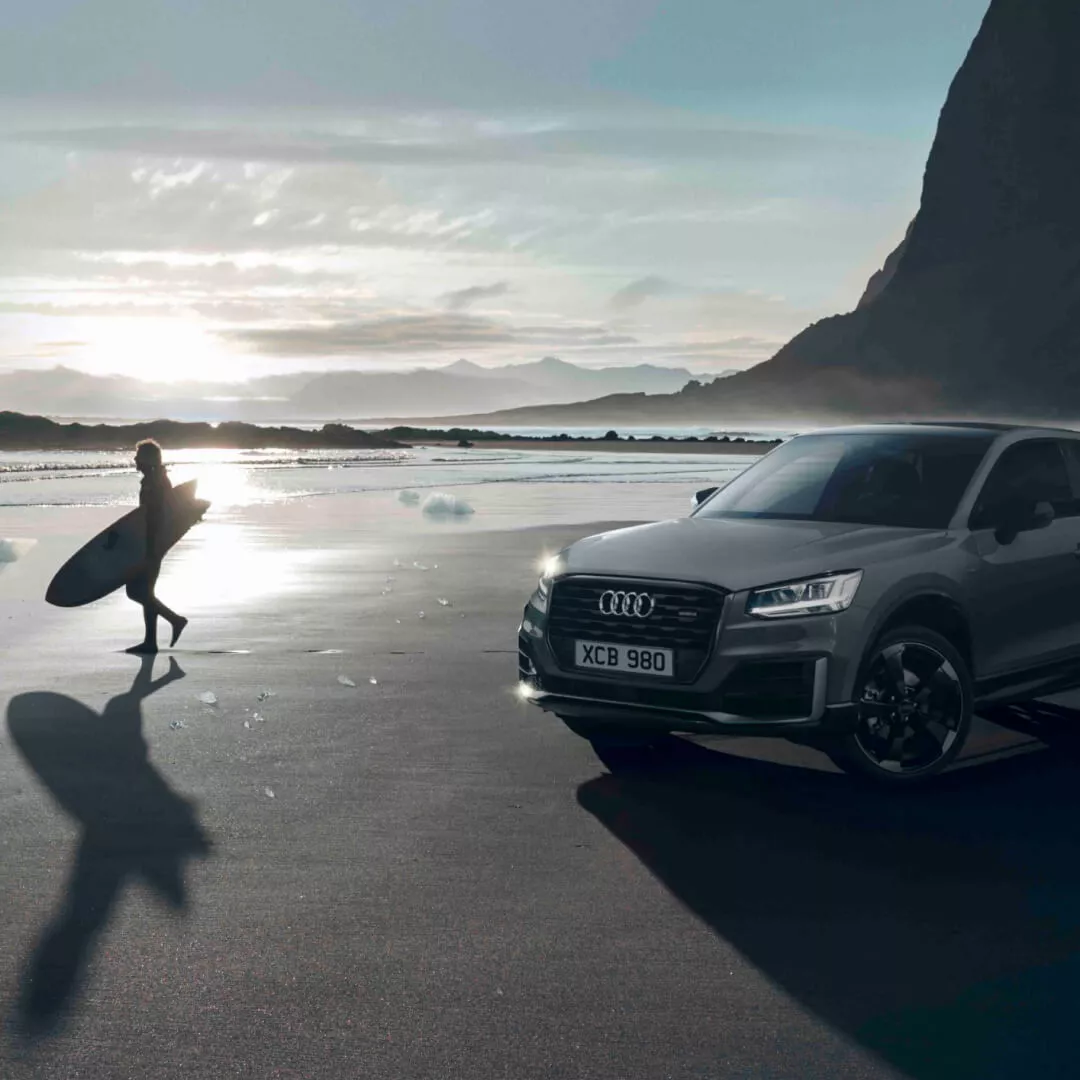 A dark grey Audi vehicle parked in a parked on beach near man walking with a surf board