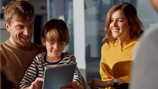 A boy holding a tablet and smiling with a mother and father beside him also smiling