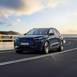 A dark blue Audi SUV drives on a coastal highway at sunset. The road curves gently with the ocean to the right and cliffs to the left. The scene is serene and dynamic.