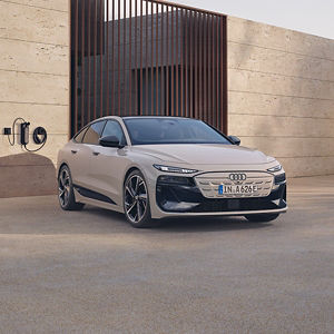 Beige Audi parked beside a modern, minimalist building with stone walls and vertical window panels. A charging station is visible nearby.
