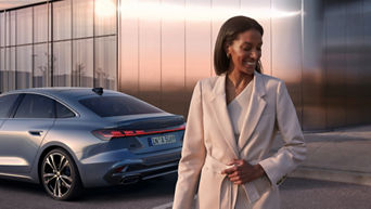 A woman in a beige suit smiles while walking in an urban setting. Behind her is a parked silver Audi car with distinctive tail lights, parked by a reflective building at sunset.