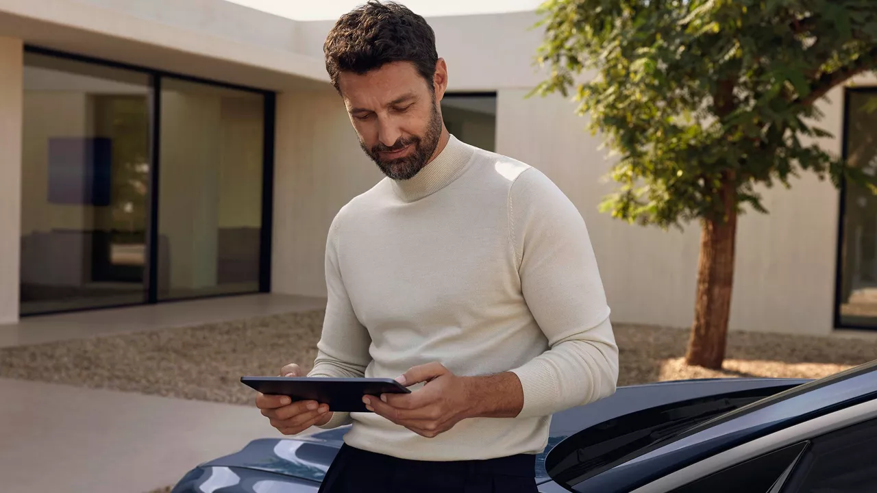 A person in a light sweater stands outdoors next to a car, holding a tablet near a modern building and tree.