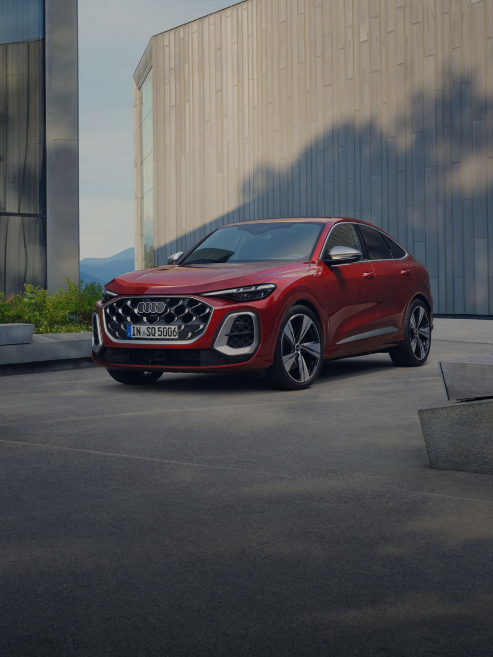 A sleek red Audi SQ5 parked in a modern urban setting with glass buildings and greenery in the background, showcasing its stylish design.