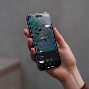 A hand holds a smartphone displaying a city map. The screen shows multiple green markers with labels, suggesting various locations or points of interest. The setting is casual, with a neutral background, conveying a sense of navigation and exploration.