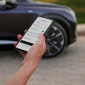 A person holds a smartphone displaying a car temperature control app next to a parked car. The setting is outdoors, creating a tech-savvy atmosphere.